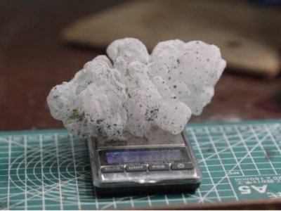 A spiky white hailstone, flecked with small dots of black paint, rests on a scale on a desk. 